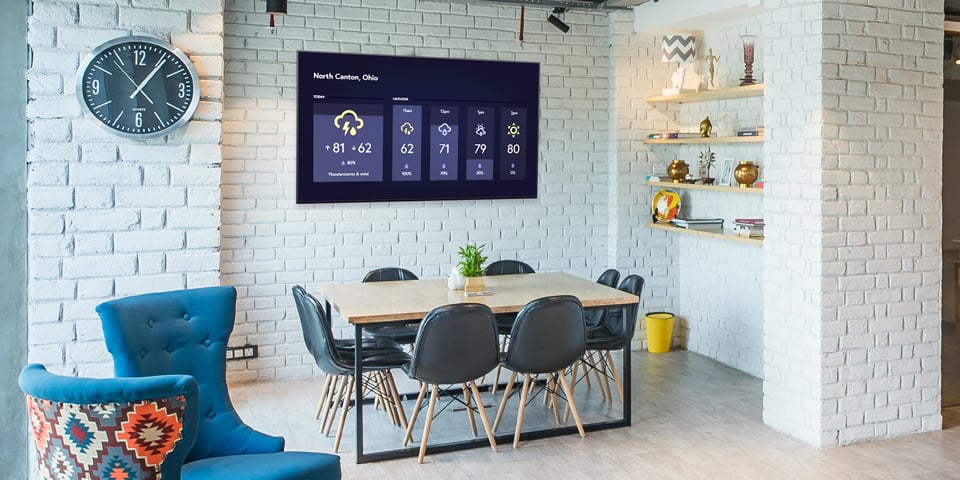 A stylish room with white brick walls and a mounted digital screen displaying a weather forecast for North Canton, Ohio above a wooden table and chairs.
