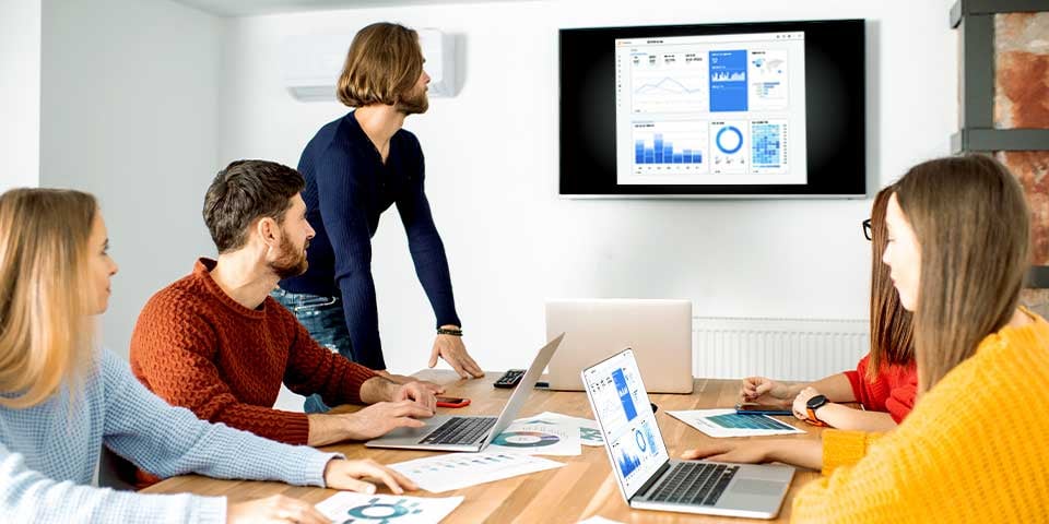 A group of five people in a meeting room viewing data dashboards on laptops and a wall-mounted screen, with one person standing and presenting.