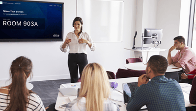 A teacher talking to students in a classroom, standing in front of a TV showing a Ditto room code 