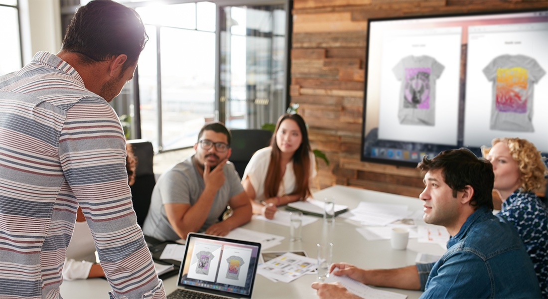 People meeting while a computer wirelessly mirrors t-shirt designs to a bigger screen